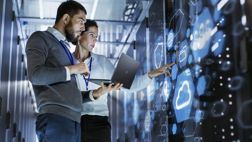 shutterstock_1062915392 1000px Male IT Specialist Holds Laptop and Discusses Work with Female Server Technician. They're Standing in Data Center, Rack Server Cabinet with Cloud Server Icon and Visualization.
